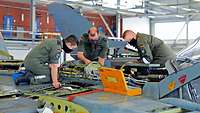 Drei Soldaten stehen auf einem Flugzeug. Alle haben den Mund-Nasen-Schutz auf