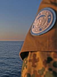 Der UNIFIL Einsatz vor der Küste des Libanon Im Vordergrund steht ein Soldat mit einem Maschinengewehr, im Hintergrund sieht man ein vorbeifahrendes Frachtschiff