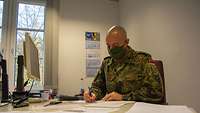 Ein Soldat mit Mundschutz sitzt an einem Schreibtisch. In der Hand hält er ein Stift. Vor ihm liegen mehrere Schriftstücke.