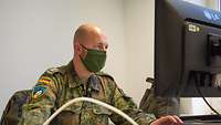 Ein Soldat mit Glatze sitzt mit einem Mundschutz vor einem PC-Bildschirm. Seine Hände liegen auf Maus und Tastatur.