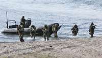 Sechs Soldaten in Flecktarn stehen mit einem Gewehr im Anschlag an einem Strand. Im Hintergrund ein Speedboot mit einem Soldaten