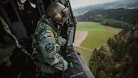 Ein Soldat hockt gesichert an der offenen Tür eines fliegenden Hubschraubers und schaut nach unten..