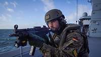 Ein Soldat in Flecktaruniform steht auf dem Deck eines grauen Schiffs und hält eine Maschinenpistole im Anschlag.