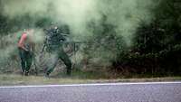 Neben einer grauen Straße stehen unbekannte Schützen hinter Büschen und schießen mit Maschinengewehren.