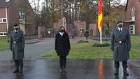 Zwischen zwei Soldaten steht eine Zivilistin, hinter ihnen an einer Gedenkstätte liegen Kränze.