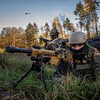 Auf einer Wiese geht ein Soldat mit einem Maschinengewehr in den Anschlag, dahinter steht ein Hubschrauber am Boden.