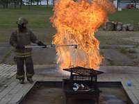 Bild 1_74 neue Brandschützer Ein Brandschützer in Schutzmontur schüttet Wasser in brennendes Fett, eine große Stichflamme entsteht.