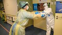 Eine Frau in Schutzkleidung reicht einer Person in Vollschutz Material an, das in einem Krankenzimmer benötigt wird