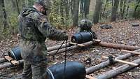 Auf dem Boden liegen Rundhölzer für die Plattform eines Tonnenfloßes. Zwei Soldaten binden vier Tonnen an die Ecken.