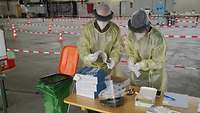Zwei Soldaten mit gelbem Schutzumhang, Gesichtsschild, Haarnetz, Maske und Handschuhen ordnen Testmaterial und Unterlagen.
