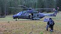 Zwei Soldaten klettern in einen Hubschrauber, der auf einer Wiese steht, davor ein Transportnetz mit Kisten.