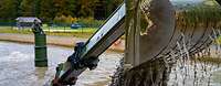 The turret and the excavator bucket of a tank protrude from a water basin. Water and mud are running out of the bucket.