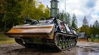 A tank with a high tube on top and a dozer blade standing in front of a lot of trees
