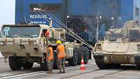 Zwei Soldaten stehen zwischen militärischen Fahrzeugen, die im Hafen vom Schiff rollen