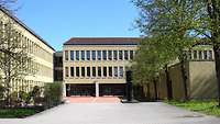 Dreigeschossiges Längs- und Quergebäude mit gelber Fassade davor ein gepflasterter Zugang.