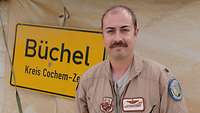 Ein Amerianischer Pilot steh vor einer Zeltwand an der ein Ortsschild aufgehangen ist mit der Aufschrift Büchel