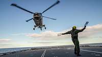 Ein Hubschrauber im Landeanflug, im Vordergrund eine Person mit Kellen in der Hand, um ihn einzuweisen
