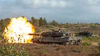 Ein Kampfpanzer schießt im Gelände. Vor der Mündung Kanone befindet sich ein Feuerball.