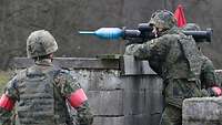 Ein Soldat steht mit einer Panzerfaust auf der Schulter hinter einer Steinwand.