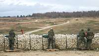 Vier Soldaten stehen mit ihren Waffen hinter einem Steinwall, dahinter ein großes Areal mit Zielscheiben.
