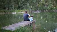 Ein Vater hält eine Angel und sitzt mit seinem Kind auf einem Steg am See.