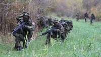Mehrere Soldaten mit Waffen und Ausrüstung knien am Rande einer Lichtung neben einer Buschgruppe und beobachten ihr Umfeld.