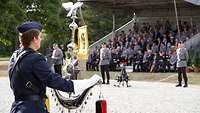 Ein Soldat mit Schellenbaum in den Händen steht vor einer Tribüne mit Zuschauern