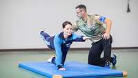 A service woman does a postural exercise on her knees. A trainer adjusts the stretched extremities.