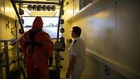 A man in an orange full-body suit stands on a treadmill in front of a monitor. The physician stands beside him.