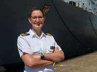 Eine schlanke, dunkelhaarige Frau mit Brille und weißem Hemd steht vor einem grauen Schiff.