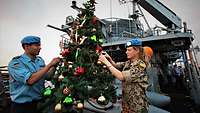 Zwei Marinesoldaten schmücken auf dem Deck eines kleinen Schiffes einen Weihnachtsbaum.