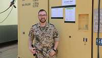 Hauptfeldwebel Wolfgang E. steht vor einem sandfarbenen Container mit einem Schild „Zahlstelle“.
