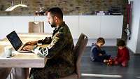 Der Vater sitzt in Uniform arbeitend an seinem Laptop während die Kinder im Hintergrund auf dem Fußboden spielen.