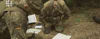 Zwei Soldaten knien auf dem Waldboden und sind über mehrere Karte gebeugt