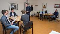 Vier Bewerber sitzen an einem Tisch und werden im Gruppensituationsverfahren geprüft. Im Hintergrund: die Prüfungskommission.