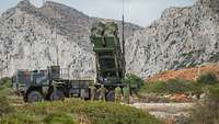 Ein Patriot-Launcher steht auf dem NATO-Schießplatz und wird von drei Soldaten aufgebaut. Dabei schauen zwei Bewerter zu.