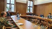 Die Frau und die Männer sitzen an einer sehr großen ovalen Tisch-Anordnung, an der man auf beiden Seiten sitzen könnte