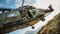 Ein grüner Hubschrauber landet auf einer Wiese am Berg. Ein Soldat schaut aus der geöffneten Seitentür auf den Boden.