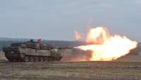Ein Kampfpanzer steht auf freier Fläche und feuert. Der Mündungsblitz ist orangerot und sehr groß.