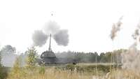 Eine Panzerhaubitze schießt, in der Luft das fliegende Geschoß, am Rohr Rauchwolken, im Vordergrund die Ähren von Schilfgras