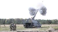Fünf Soldaten stehen hinter einer Panzerhaubitze und ziehen an einem Seil, ein Geschoss fliegt durch die Luft 