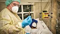 A man in full protective gear holds an hourglass and measures the pulse of the patient lying in bed.