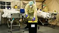 A man in full protective gear is standing in front of two patient's beds and throws something into a medical waste bin.