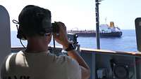 Ein Boardingsoldat der Fregatte „Hamburg“ mit Headset und Fernglas in der Hand beobachtet das andere Schiff