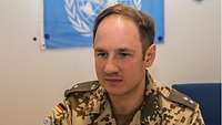 Ein Soldat sitzt an seinem Arbeitsplatz, im Hintergrund hängt die Flagge der Vereinten Nationen 
