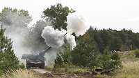 Eine Panzerhaubitze schießt, Rauchwolken umgeben das Rohr, rechts ein aufsteigendes Geschoß 