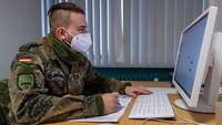 Ein Soldat mit Mund-Nasenschutz sitzt an einem Schreibtisch und schaut auf den Bildschirm eines Computers.