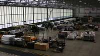 Blick von oben in eine große Lagerhalle mit Transportbehältern, verpackten Geräten, Gabelstaplern und einigen Soldaten.