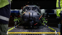 Ein Militärhubschrauber ohne Rotorblätter wird an Bord eines Transportflugzeuges gezogen.