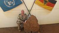 Ein Soldat kniet an einem Stein vor der Deutschland- und der VN-Flagge, im Hintergrund ein grüner Stofffrosch von seiner Tochter